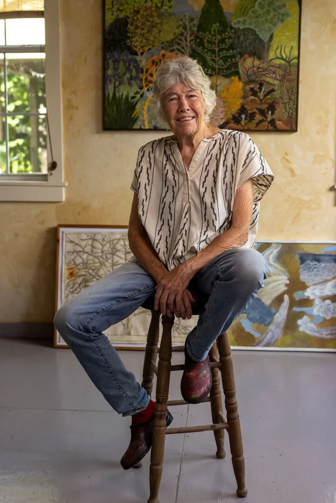 An older woman with gray hair, wearing a patterned short-sleeve shirt, jeans, and red shoes, sits on a wooden stool in a sunlit room with abstract artwork on the walls behind her.