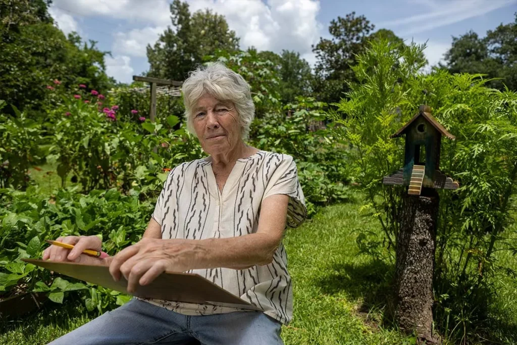 An older person with gray hair sits in a lush garden, holding a clipboard and pencil, appearing to sketch or write. Green plants and a small birdhouse on a tree stump are visible nearby under a sunny sky.