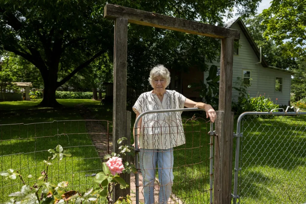 An older woman stands behind a metal gate in a garden, surrounded by greenery and flowers, with a house and large tree in the background on a sunny day.