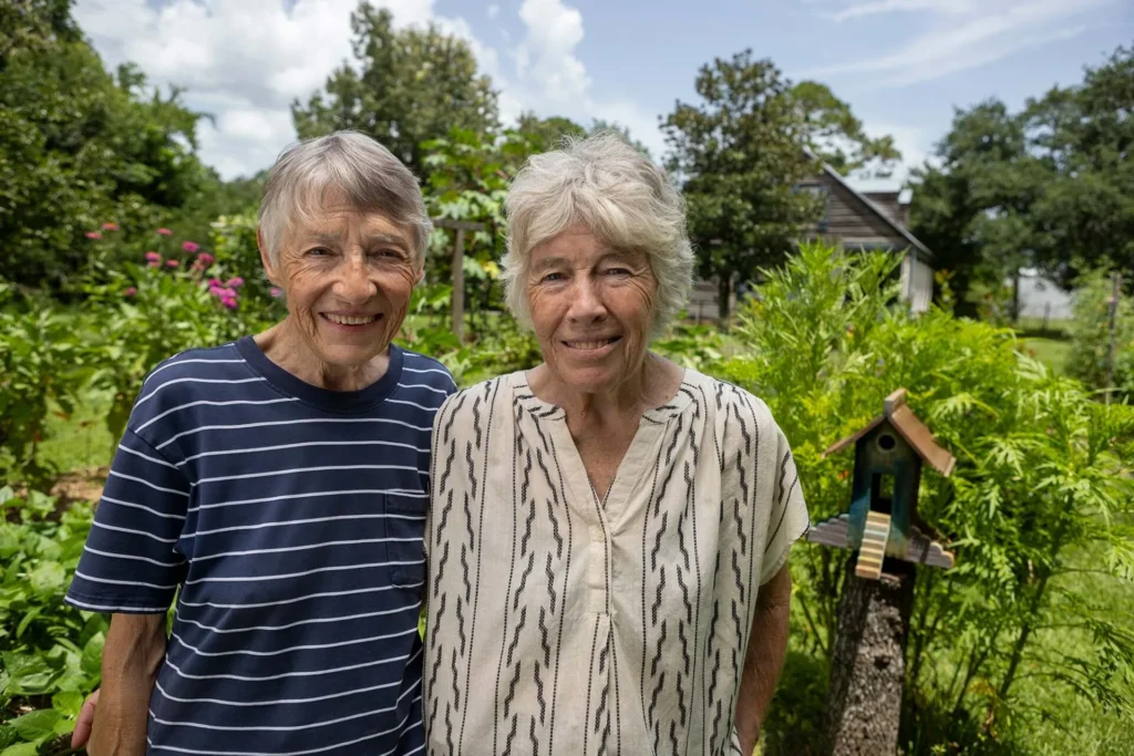 Two older women stand closely together, smiling in a lush, green garden with flowers and trees. A small wooden birdhouse is visible beside them, and a house appears in the background under a partly cloudy sky.
