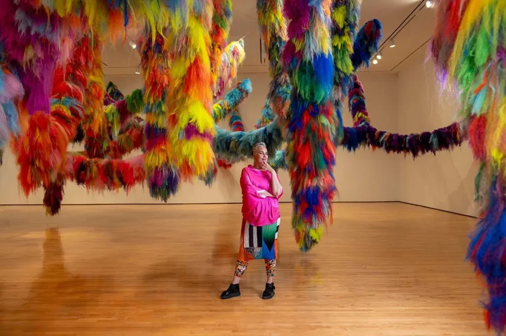 A person in colorful clothing stands in a gallery beneath large, hanging, multicolored fuzzy installations that stretch across the room, creating an immersive, whimsical environment.