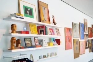 White wall with three floating shelves displaying books, postcards, small art pieces, wooden busts, and decorative objects. Framed artworks are hung above and beside the shelves, creating a colorful, creative display.