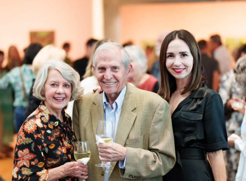 Three adults pose and smile at a social event, holding glasses of wine. The woman on the left wears a floral dress, the man in the center wears a tan suit, and the woman on the right wears a black dress. A crowd is in the background.