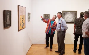 A group of people observe and discuss artwork displayed on the walls of a gallery. One woman in a colorful shirt points at a painting while talking to a man in a tie. Others view the art quietly.