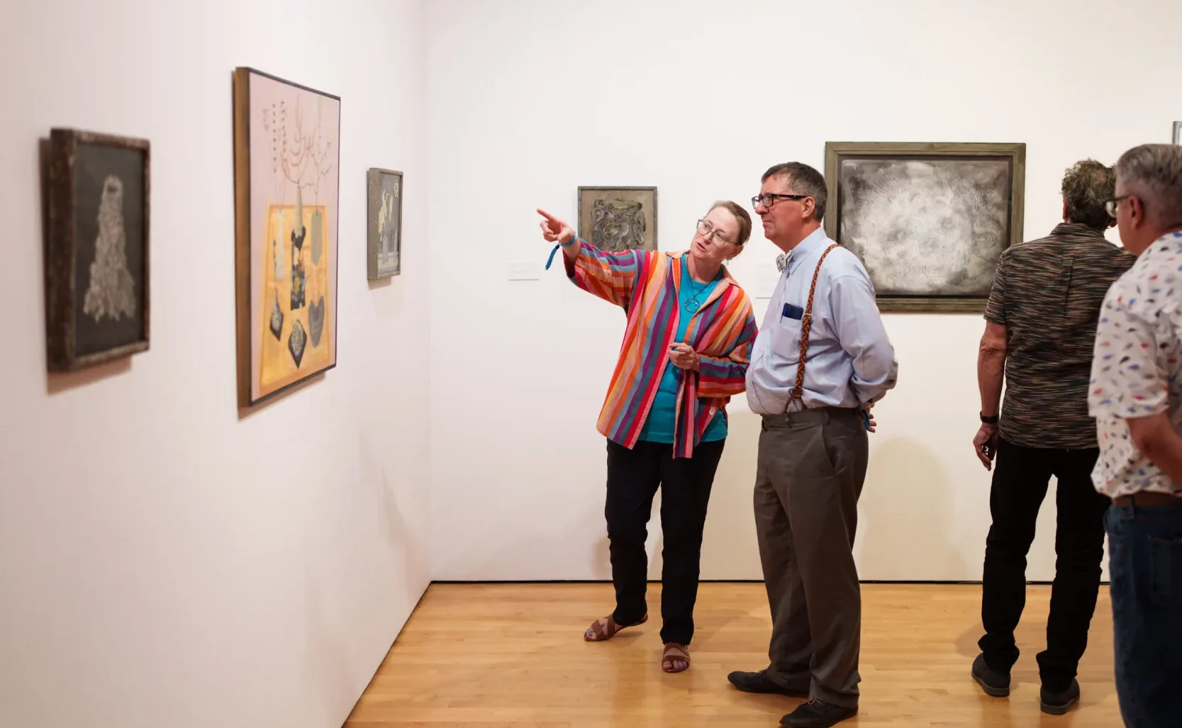 A group of people observe and discuss artwork displayed on the walls of a gallery. One woman in a colorful shirt points at a painting while talking to a man in a tie. Others view the art quietly.