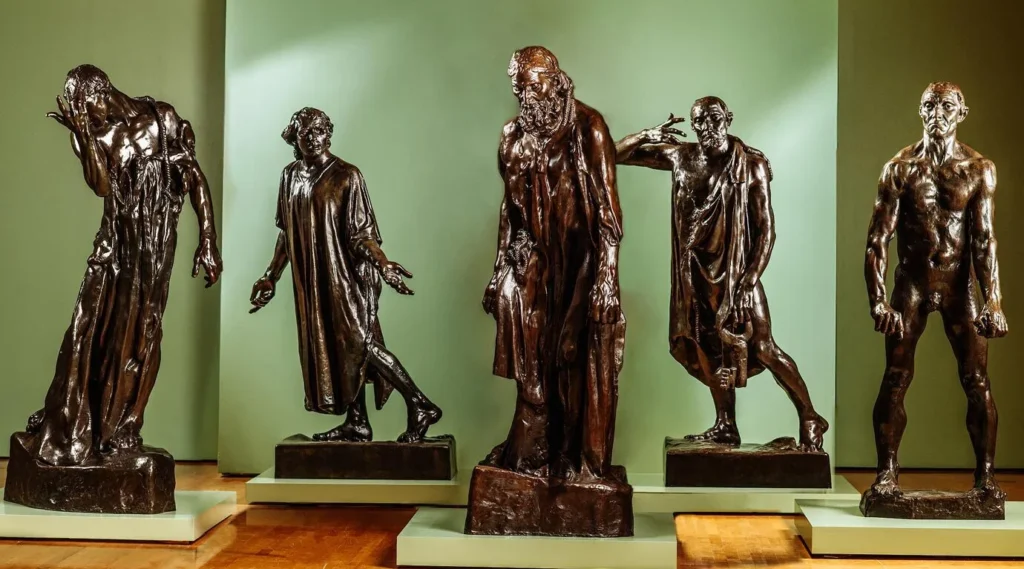 Five bronze statues of men in dramatic, anguished poses stand on individual platforms against a green brisco background. The life-sized, detailed figures are arranged in a staggered formation, capturing intense emotion and presence.