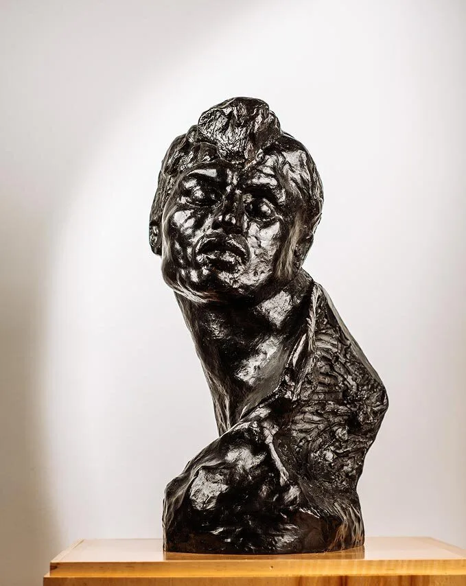 A dark, glossy bronze brisco sculpture of a human torso and head, with a dramatic, expressive face and textured surface, is displayed on a wooden base against a plain light background.