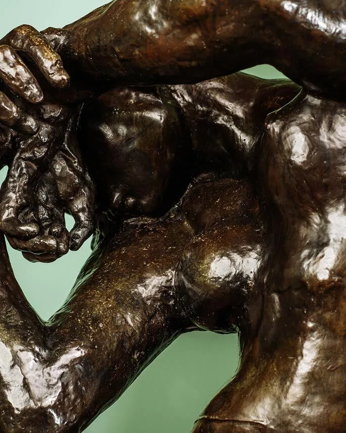 Close-up of a bronze Brisco sculpture depicting two intertwined human figures, with one figure holding the other’s hand over their head, set against a soft green background.