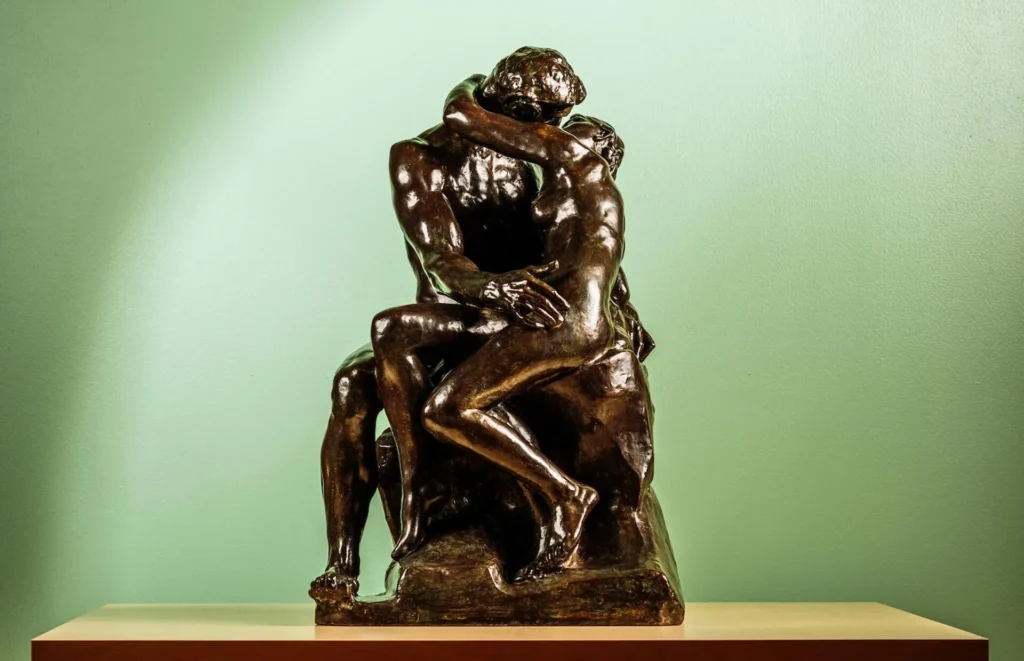 A bronze Brisco sculpture of two nude figures embracing and kissing, seated closely together on a pedestal against a plain light green background.