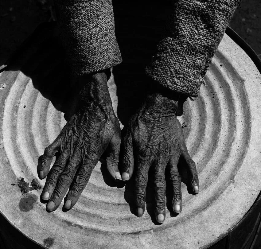 Close-up of two elderly hands with visible wrinkles and veins, resting flat on a circular, ridged brisco surface. The person is wearing a textured, long-sleeved garment. The image is in black and white.