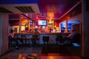 A dimly lit bar with a few people sitting on stools at the counter. The bar area is illuminated with warm orange and cool blue lighting. Bottles and a TV are visible behind the counter.