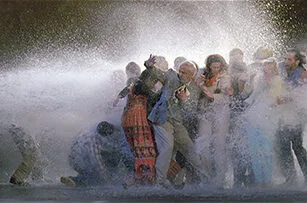 A group of people, dressed in formal and casual clothing, huddle together as they are sprayed with water from a strong jet, creating a dramatic splash and mist around them.