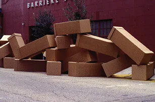 Several large, rectangular cardboard boxes are stacked haphazardly and spilling onto a street in front of a red building with windows and trees in the background.