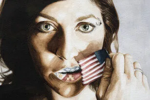 A woman holds a small American flag over her mouth, partially covering her lips, which show a tattoo-like pattern. She has light eyes, brown hair, and wears a ring on her finger.