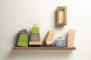 A wooden shelf holds several abstract, geometric wooden blocks painted in green, blue, and natural tones. Above the shelf, a small empty wooden box is mounted on a white wall.