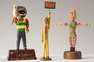 Three colorful carved wooden figurines stand side by side: a man with an alligator head holding a chicken, a man in a yellow suit holding a sign, and a woman in a green bikini with tattoos and outstretched arms.