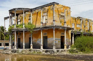 A large, weathered yellow building with peeling paint, broken windows, and overgrown plants. The structure appears abandoned and partially damaged, with columns supporting a second-floor balcony.