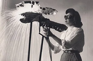 A woman wearing sunglasses and a white blouse uses a welding tool to work on a metal sculpture, with sparks flying as she welds. The scene is in black and white.