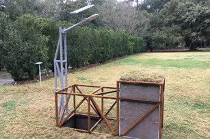 A wooden and metal composting structure with an open top stands on a grassy lawn. There is a solar-powered lamp attached to a pole beside it, and trees and bushes are in the background.