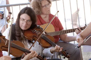 Three people play string instruments together, including two fiddles and a guitar, in an outdoor setting. A woman with long dark hair is focused on playing the fiddle in the foreground.