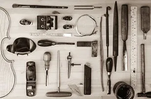 A variety of violin-making tools, including files, screwdrivers, a plane, rulers, clamps, and pieces of wood, neatly arranged on a wooden surface alongside a partial violin outline.