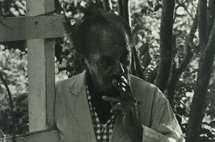 Black-and-white photo of a person in a light jacket and checkered shirt standing outdoors, partially in shadow, holding and smoking a cigarette near their mouth with trees in the background.