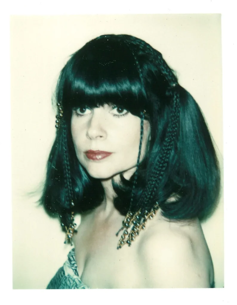 A woman with dark, shoulder-length hair styled with braids and gold beads looks at the camera. She has bangs, striking eye makeup, and red lipstick, wearing a strapless patterned top against a light background.