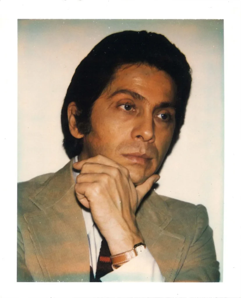 A man in a tan suit and striped tie rests his chin on his hand, gazing thoughtfully to the side. He has dark hair and wears a wristwatch. The photo has a Polaroid-style frame.