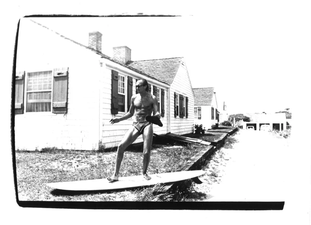 A man in swim trunks stands on a surfboard on the grass in front of a row of beach cottages, holding what appears to be a leash and gesturing with his hands, as if balancing. The scene is in black and white.