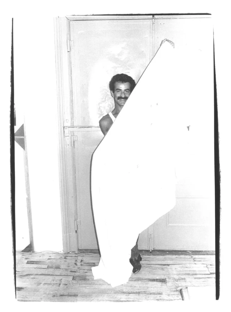 A man with a mustache stands indoors on a wooden floor, partially hidden behind a large white sheet he is holding, with a closed door in the background.