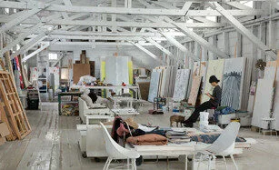 A spacious, white-painted art studio with high ceilings, scattered sofas, and various large canvases. Several artists work or relax among art supplies and easels in a creative, informal setting.