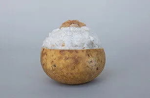 A brown fruit shell, possibly a bael, filled with crushed ice and topped with brown sugar, set against a plain gray background.