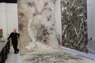 A person stands beside large abstract artworks hanging in a spacious studio. The artworks feature intricate black, white, and gray patterns. The person is dressed in black and is leaning against a table.