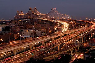 A city bridge and highway interchange at night with heavy traffic; streaks of car lights are visible, and the bridge is illuminated by rows of bright lights.