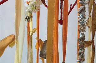 Various ribbons and fabric strips in shades of yellow, orange, and gold hang vertically. Some are adorned with white flowers, metallic elements, and a small bell against a light background.