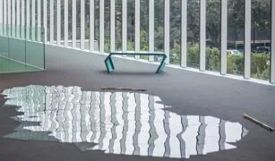 A puddle of water on a dark floor reflects tall vertical windows; a modern, angled bench sits nearby inside a bright, glass-walled building.