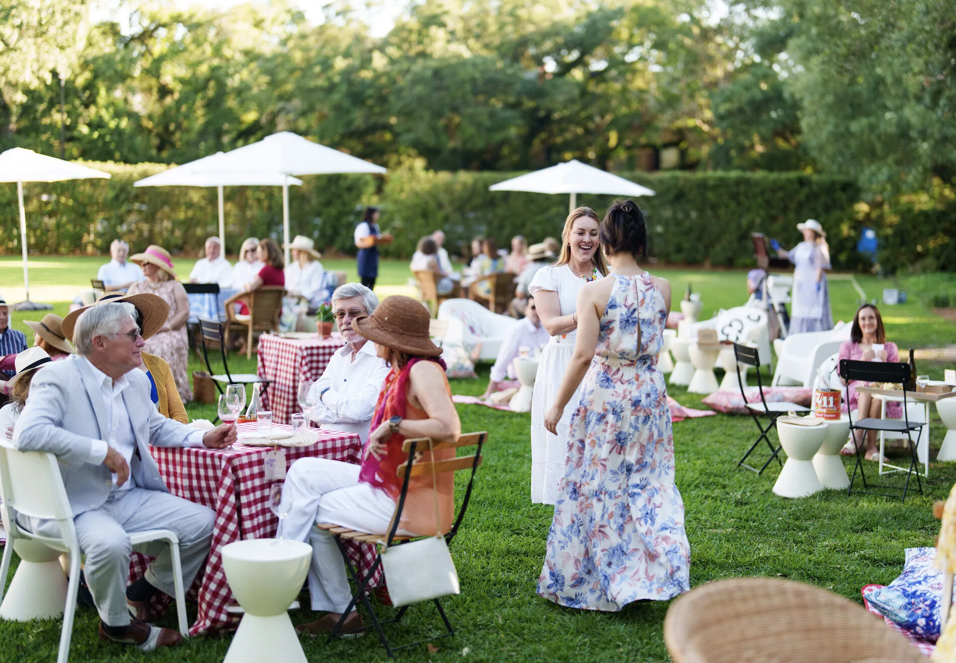 People dressed in summer attire sit and stand around tables with red checkered tablecloths at an outdoor garden party on a sunny day. White umbrellas provide shade, and trees are visible in the background.