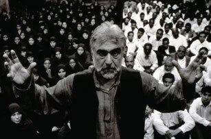 A man with gray hair and a beard stands with raised hands in front of a large seated crowd, mostly dressed in dark or white clothing, in a black-and-white photograph.