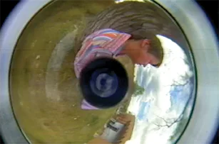 A distorted, circular fisheye view shows a person outdoors near a tree. The person appears to be wearing a colorful striped shirt, and the scene includes grassy ground and partly cloudy sky.