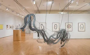 A modern art gallery with hardwood floors features a large, suspended wire sculpture resembling flowing waves. Framed abstract drawings hang on white walls in the background. Spotlights illuminate the space.