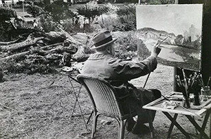 An older man wearing a hat and coat sits in a wicker chair outdoors, painting a landscape on a canvas set on an easel. Art supplies are on a small table nearby, and trees and foliage fill the background.