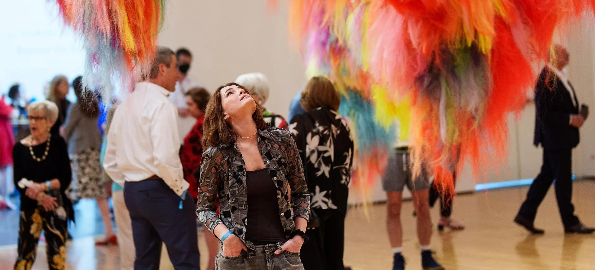 A woman looks up at colorful, fluffy art installations hanging from the ceiling, surrounded by people mingling in a bright gallery space.