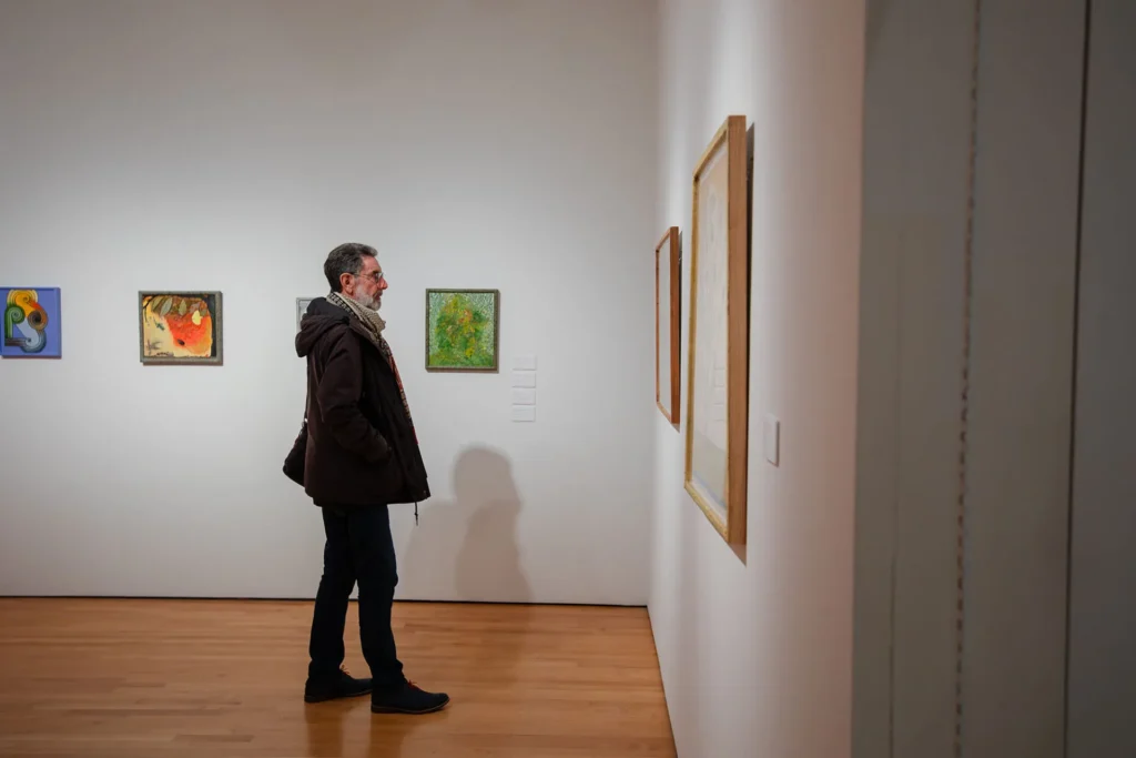 A man wearing a coat and scarf stands in an art gallery, looking at a framed artwork on a white wall. Several colorful paintings are displayed behind him. The floor is wooden.