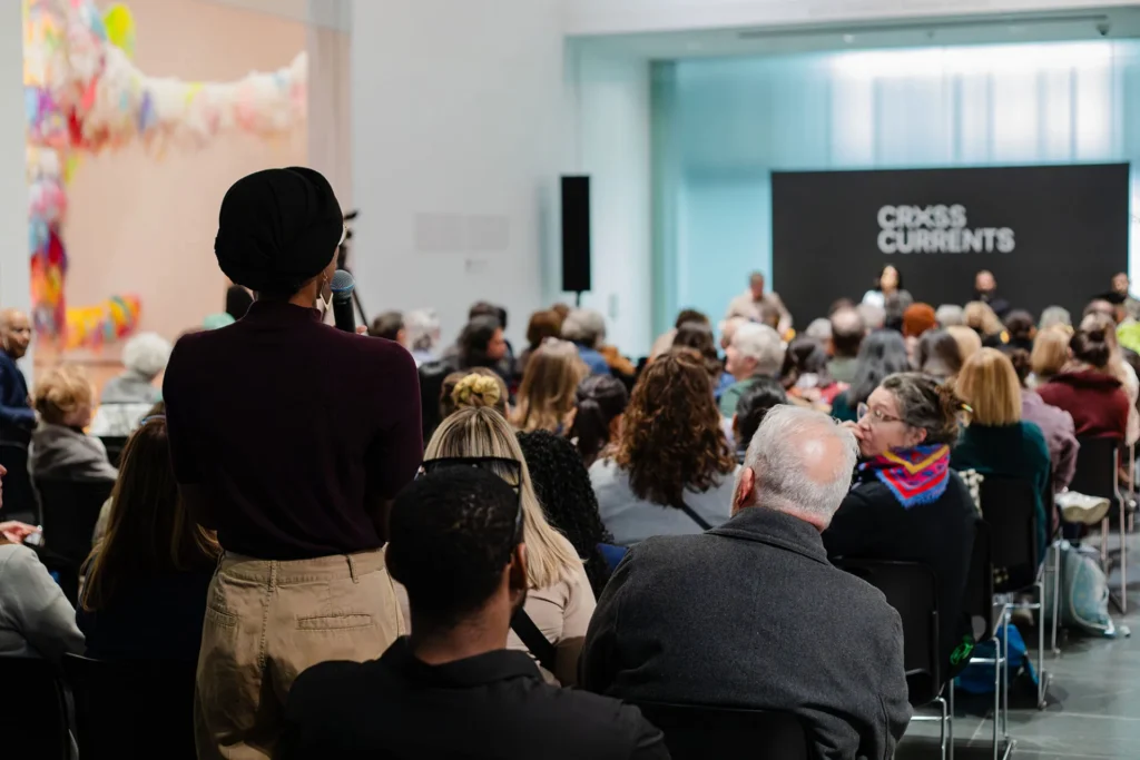 A person stands with a microphone, addressing a seated audience at an indoor event. The room is modern with large art on the wall and a stage in the background displaying the words Cross Currents.