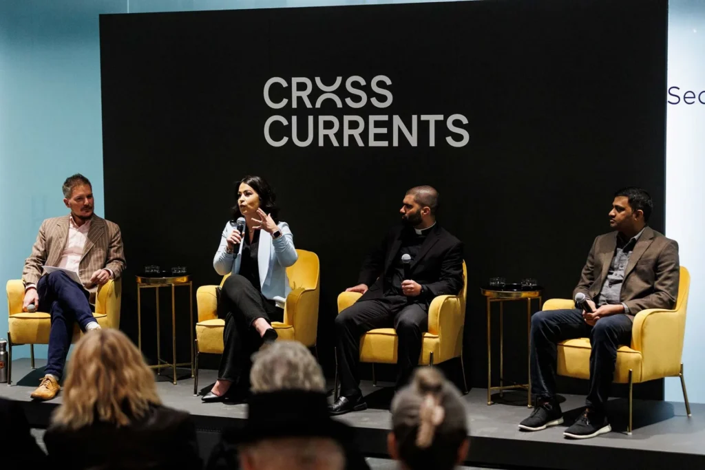Four people sit on yellow chairs on a stage, engaged in a panel discussion under a sign that reads “Cross Currents.” Audience members are seated in the foreground, listening attentively.