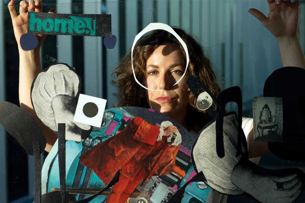 A woman stands behind a glass panel decorated with colorful cut-out shapes, photos, and the word home! Her face is framed by a white circle, and sunlight casts strong shadows on her and the art.