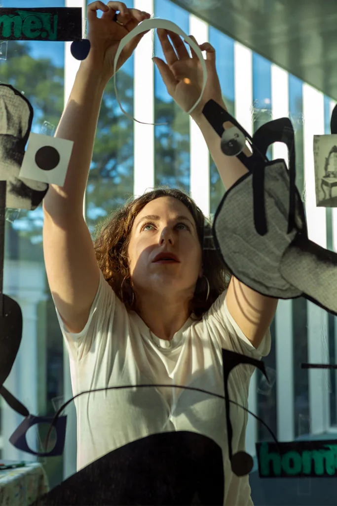 A woman in a white shirt reaches up to place or adjust circular and abstract cutout shapes on a glass window, with sunlight streaming in and trees visible outside.
