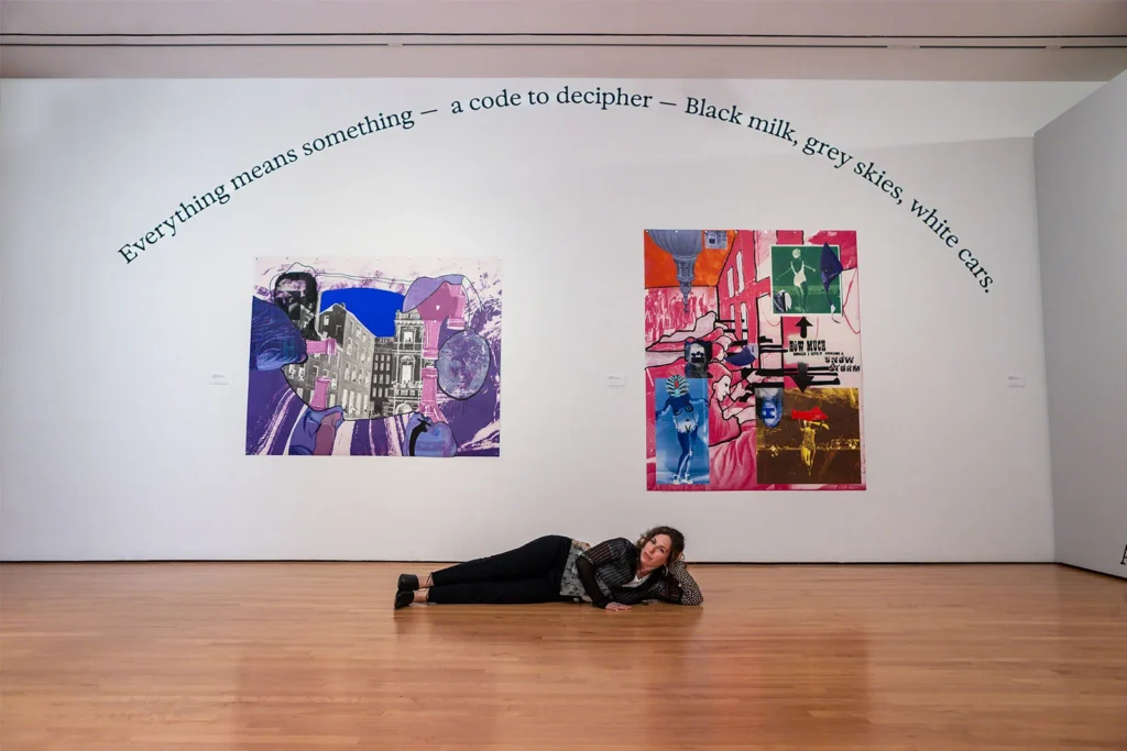 A woman lies on a wooden floor in front of a white wall displaying two colorful abstract paintings. Above the paintings, text on the wall reads: Everything means something—a code to decipher—Black milk, grey skies, white cars.