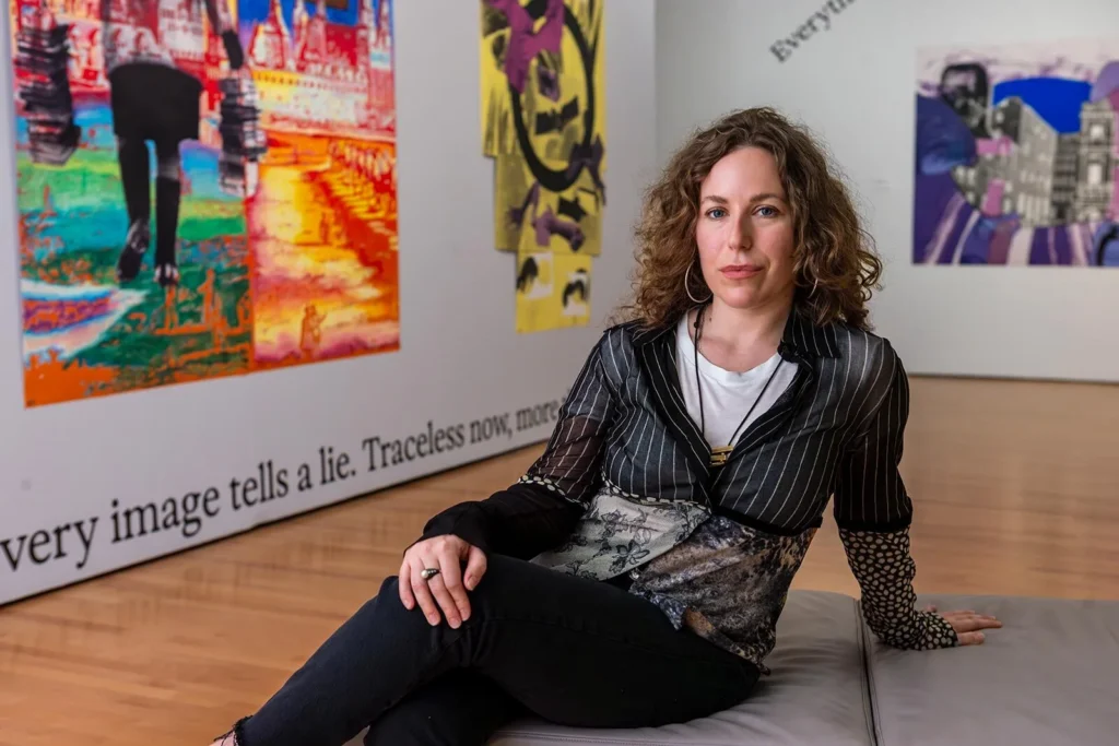 Mare Martin, a woman with curly hair, sits on a bench in an art gallery with colorful abstract paintings and text on the walls behind her. She faces the camera, wearing a patterned blouse and black pants.