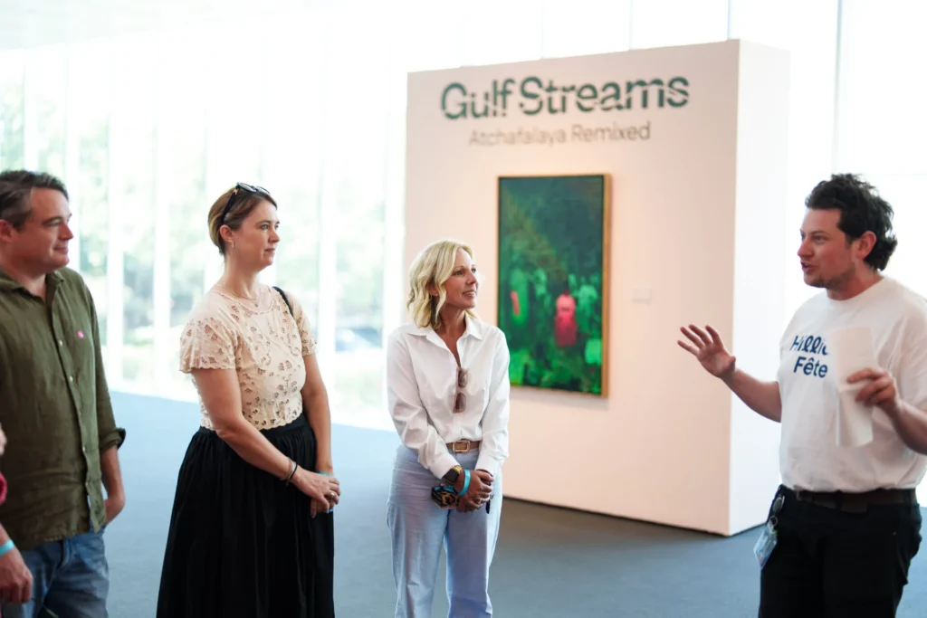 Four people stand in a gallery, three listening to a man speaking. Behind them is a sign reading Gulf Streams: Atchafalaya Remixed and a painting with green tones on a white wall.